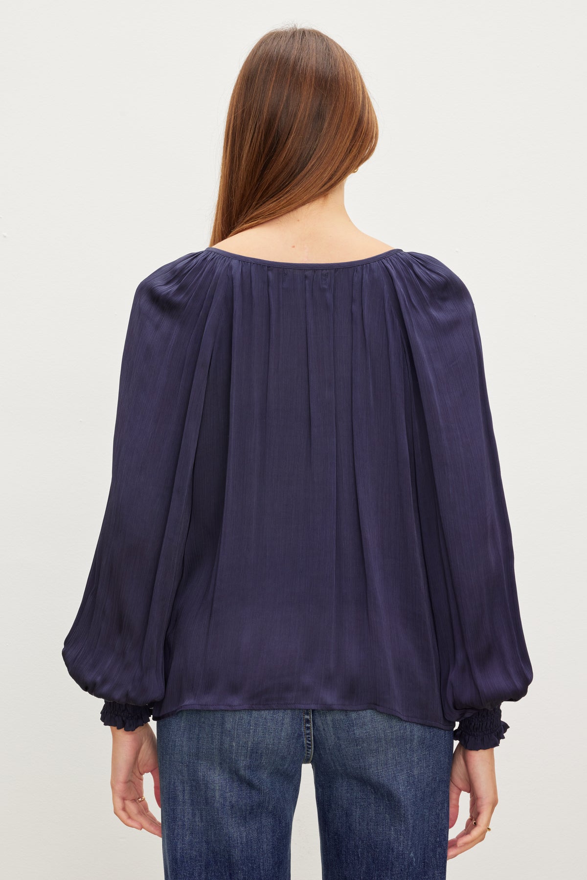 A person with long hair wearing a navy blue Velvet by Graham & Spencer HELEN TOP with puffy sleeves and blue jeans, facing away from the camera.-37073685086401