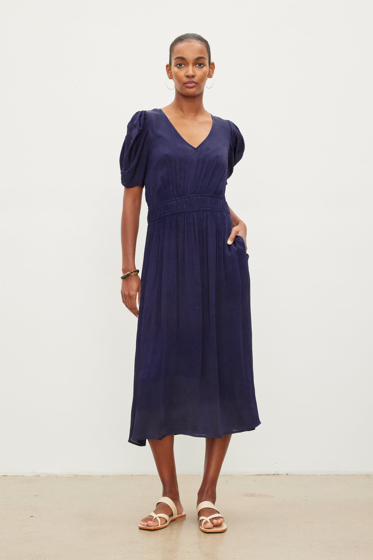   A person stands against a plain white background. They are wearing a navy blue JOSEY DRESS by Velvet by Graham & Spencer with short puff sleeves and a fitted waistband. They have hoop earrings and white sandals. 