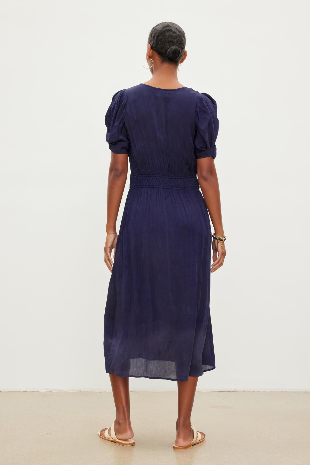   A person with short hair stands facing away from the camera, wearing a dark blue, knee-length JOSEY DRESS with puff sleeves and sandals by Velvet by Graham & Spencer. 