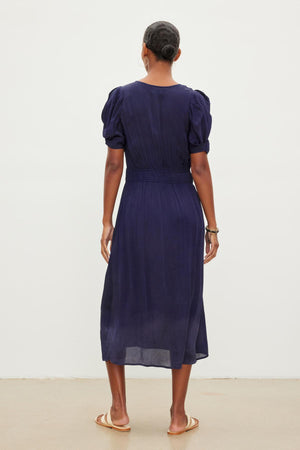 A person with short hair stands facing away from the camera, wearing a dark blue, knee-length JOSEY DRESS with puff sleeves and sandals by Velvet by Graham & Spencer.