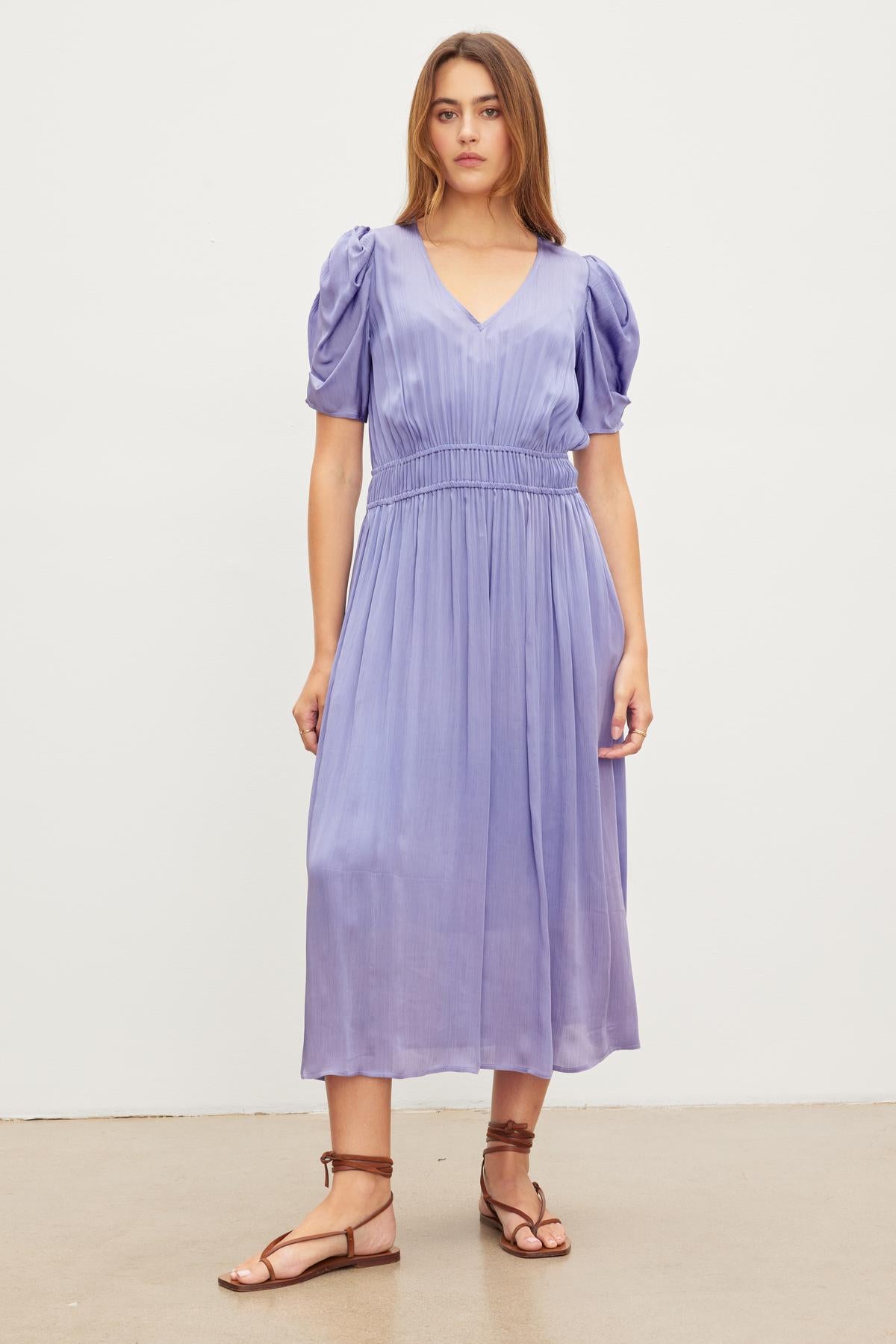   A woman stands in front of a white background wearing the Velvet by Graham & Spencer JOSEY DRESS, a light purple knee-length dress with puffed sleeves, and brown sandals. 