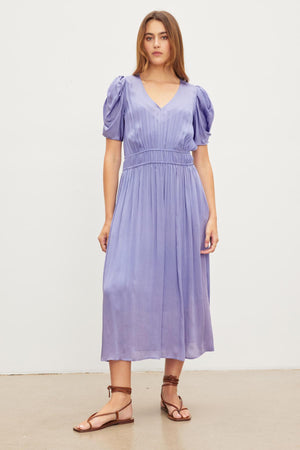 A woman stands in front of a white background wearing the Velvet by Graham & Spencer JOSEY DRESS, a light purple knee-length dress with puffed sleeves, and brown sandals.