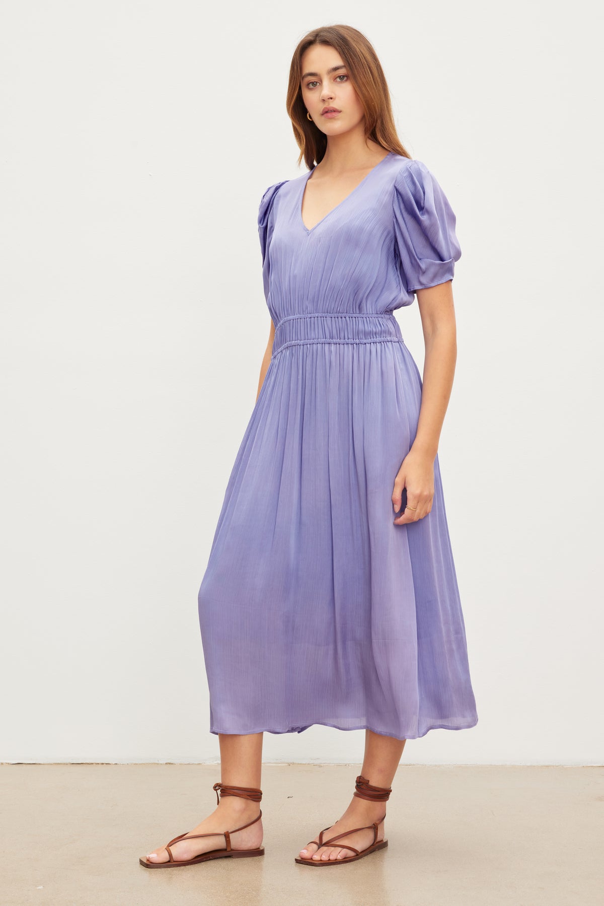 A woman stands wearing a light purple, knee-length JOSEY DRESS by Velvet by Graham & Spencer with short puffed sleeves. She is also wearing brown strappy sandals. The background is plain white.-37073688658113