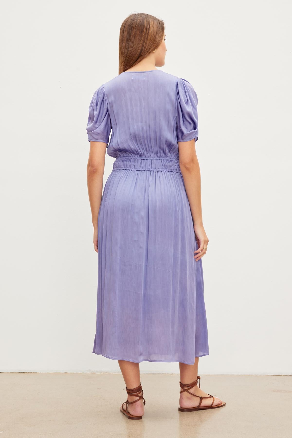   A woman is standing with her back facing the camera, wearing a mid-length, short-sleeve, purple JOSEY DRESS by Velvet by Graham & Spencer and brown sandals against a plain white background. 