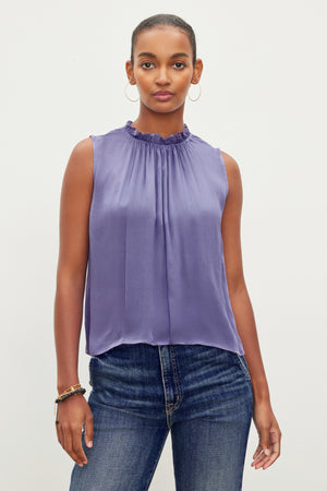 A person stands against a plain background, wearing the Velvet by Graham & Spencer KIANA RUFFLE NECK TANK TOP and blue jeans.