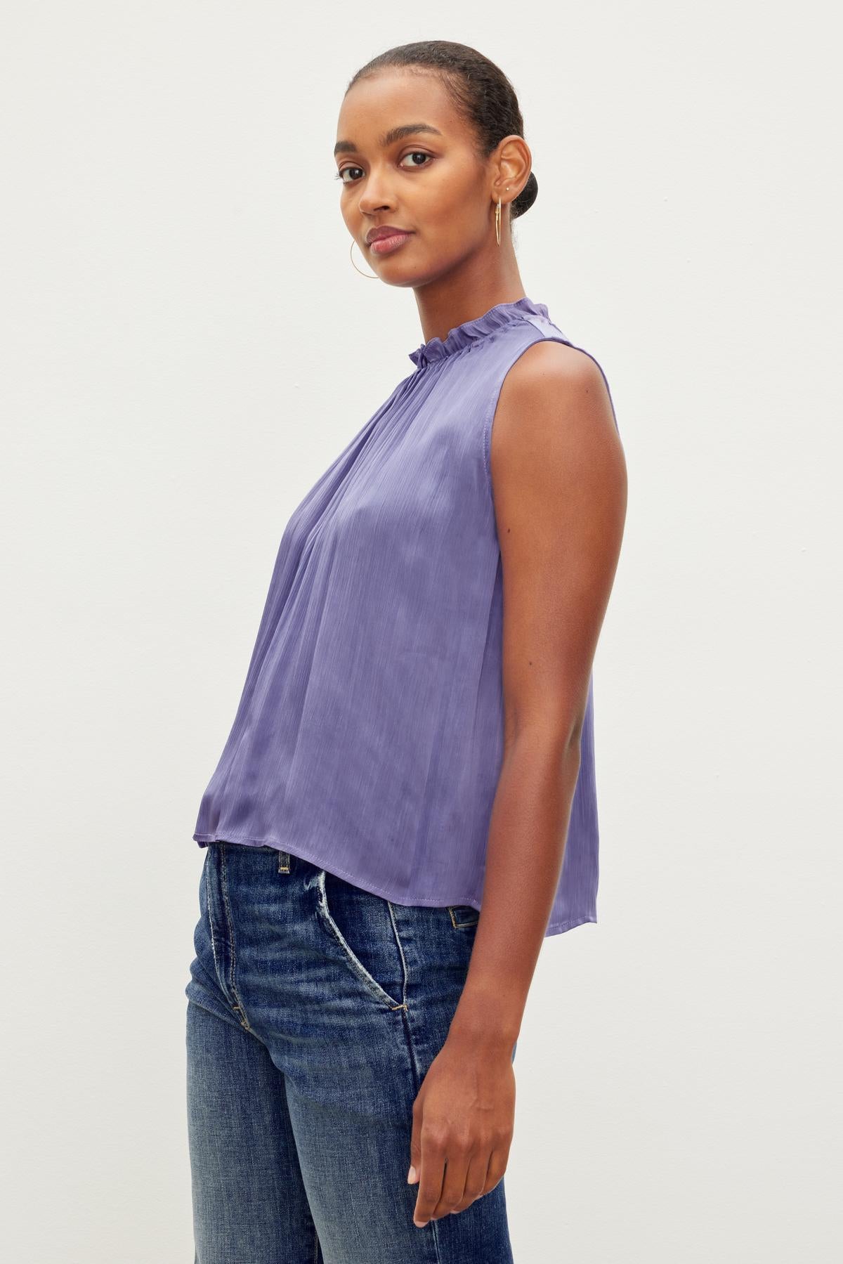   Person standing sideways wearing a sleeveless purple KIANA RUFFLE NECK TANK TOP by Velvet by Graham & Spencer and blue jeans, looking forward with a neutral expression. 