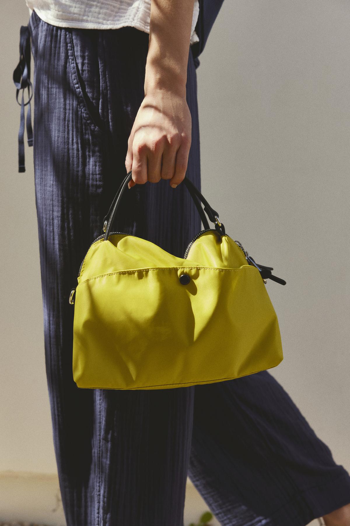 A person holding a Velvet by Graham & Spencer NYLON CROSSBODY BAG, against a plain background. The person is wearing textured blue pants and a partially visible white top.-36443575648449