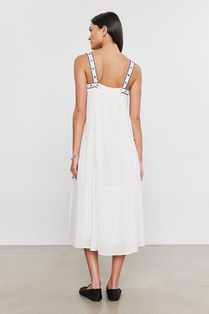 Woman in a white mid dress with decorative straps, standing facing away from the camera, on a plain background wearing the Riley dress by Velvet by Graham & Spencer.