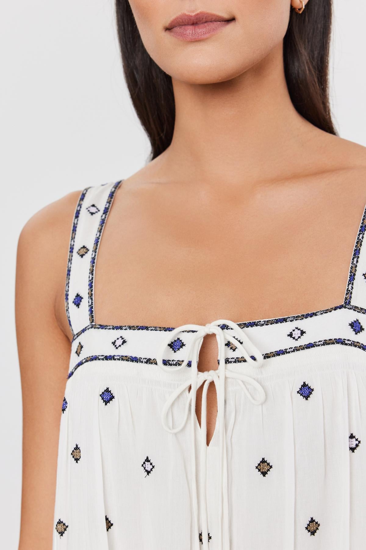   Close-up of a woman wearing a white sleeveless Velvet by Graham & Spencer RILEY DRESS with blue and black embroidered details and a tie at the neckline. 