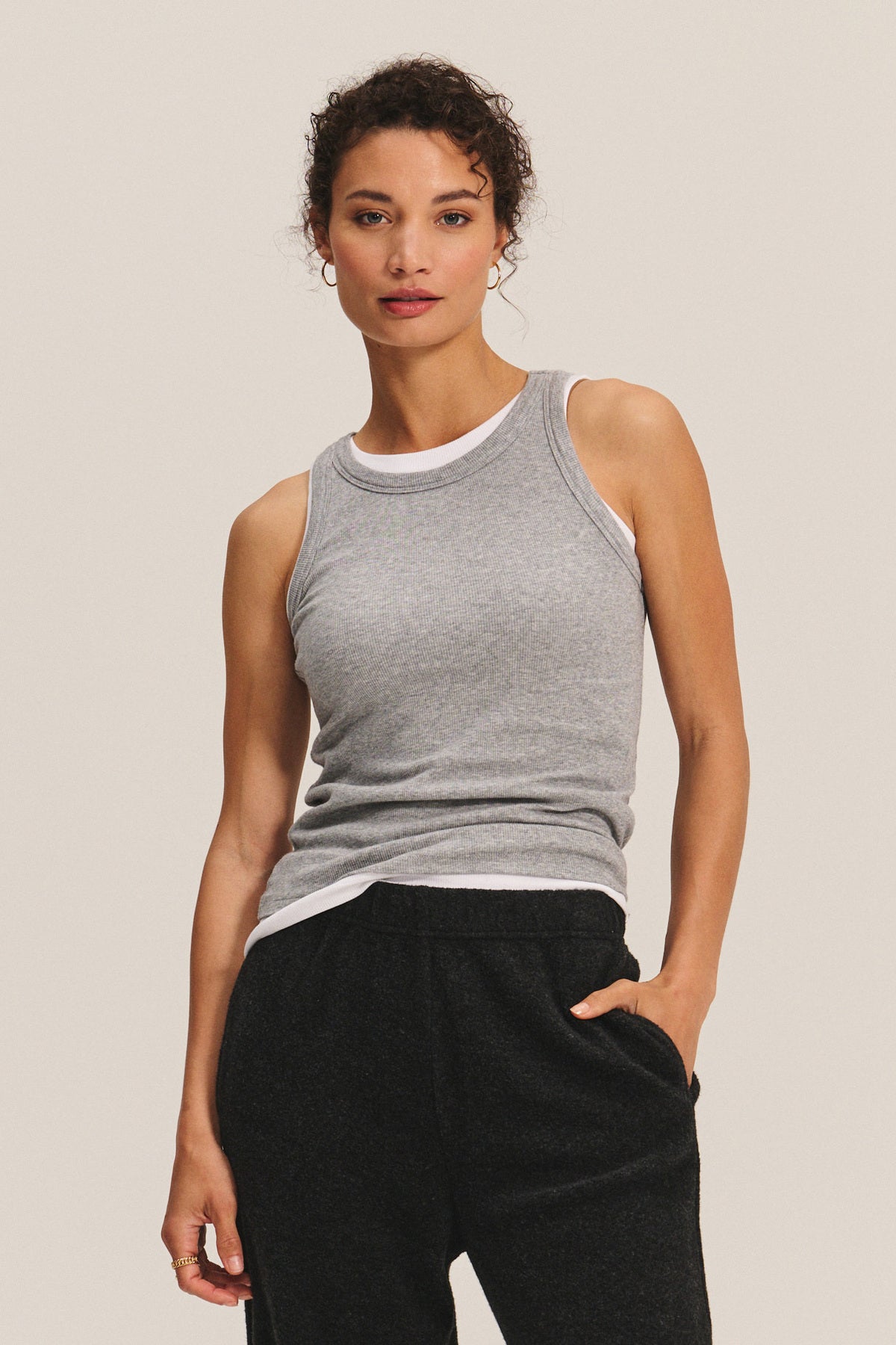 A woman stands against a plain light background wearing the CRUZ TANK TOP by Velvet by Jenny Graham in grey, layered over a white ribbed tank with black pants—an effortlessly versatile look for any capsule wardrobe.