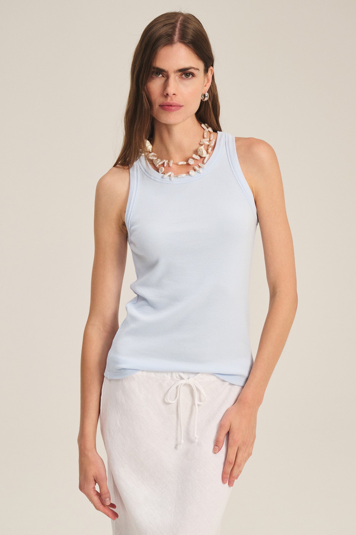 Wearing the CRUZ TANK TOP by Velvet by Jenny Graham, a white drawstring skirt, and a chunky shell necklace, she stands against a plain background—perfect capsule wardrobe inspiration.