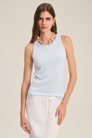Wearing the CRUZ TANK TOP by Velvet by Jenny Graham, a white drawstring skirt, and a chunky shell necklace, she stands against a plain background—perfect capsule wardrobe inspiration.