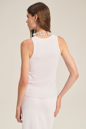 Woman stands with her back to the camera in the CRUZ TANK TOP by Velvet by Jenny Graham, a white cotton-modal blend ribbed tank—styled with a white skirt, shell necklace, and earrings for a timeless capsule wardrobe look.