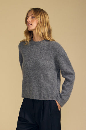A person wearing the Velvet by Graham & Spencer JUNE SWEATER—a loose gray wool-cashmere knit—paired with dark pants, stands against a plain beige background, gazing slightly to the side.