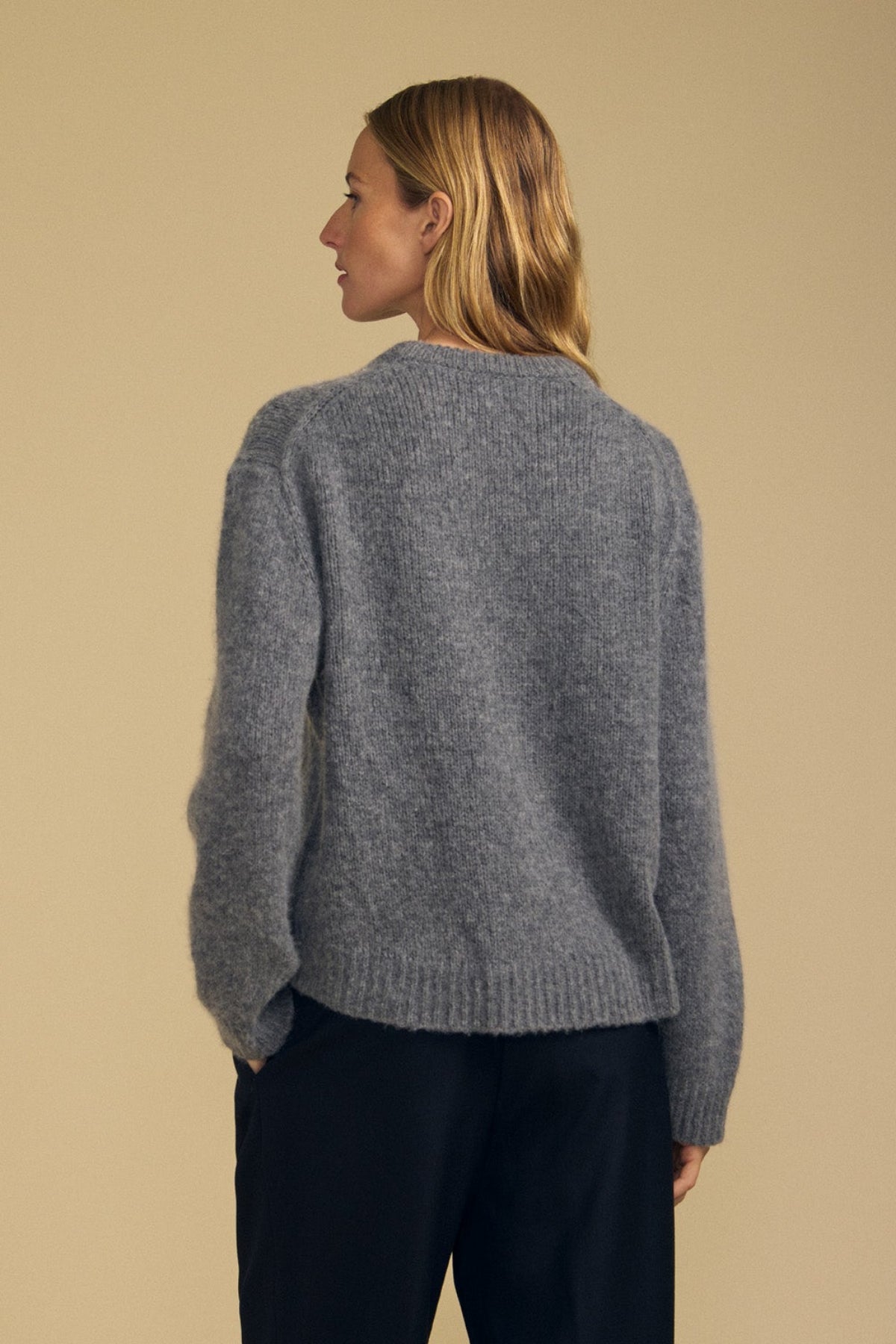 A person with light brown hair wears the JUNE SWEATER by Velvet by Graham & Spencer—a loose wool-cashmere sweater in gray—paired with dark pants, standing with their back to the camera against a beige background.