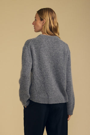 A person with light brown hair wears the JUNE SWEATER by Velvet by Graham & Spencer—a loose wool-cashmere sweater in gray—paired with dark pants, standing with their back to the camera against a beige background.