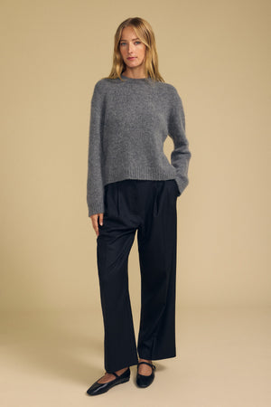 A woman stands against a beige background wearing a modern silhouette: the JUNE SWEATER by Velvet by Graham & Spencer in cozy grey wool-cashmere, black wide-leg trousers, and black Mary Jane shoes.