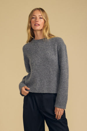 A person with long blonde hair wears the JUNE SWEATER by Velvet by Graham & Spencer—a cozy wool-cashmere piece—paired with black pants, standing against a plain beige background.