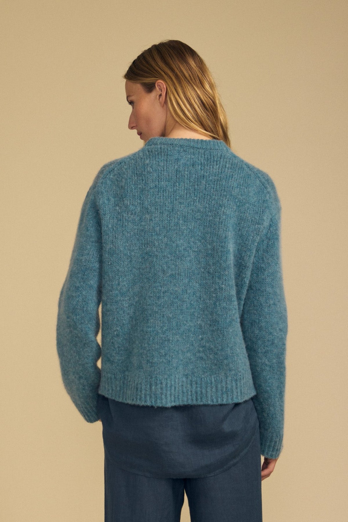 A person with long blonde hair, seen from behind, wears the JUNE SWEATER by Velvet by Graham & Spencer—a teal wool-cashmere piece with a modern silhouette—layered over a blue shirt, standing against a beige background.-41964563038401