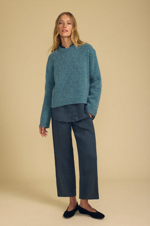 A woman stands against a beige background wearing the Velvet by Graham & Spencer JUNE SWEATER in teal over a dark gray shirt, paired with wide-leg pants and black shoes for a modern silhouette.