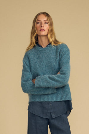 A person with long blonde hair stands with arms crossed against a beige background, wearing the Velvet by Graham & Spencer JUNE SWEATER—a cozy blue knit wool-cashmere top—layered over a dark blue shirt and matching pants.