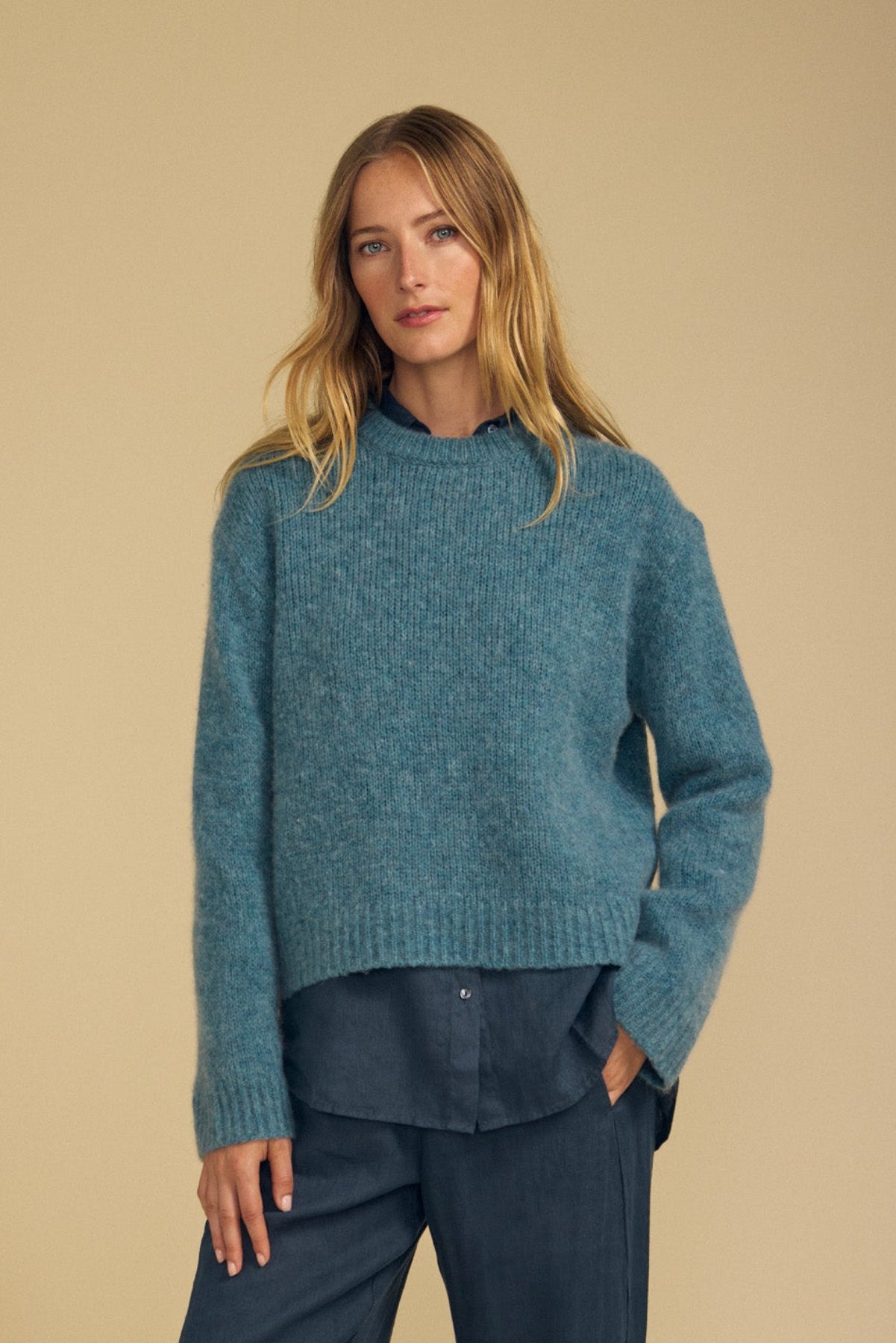 A person with long blonde hair wears the JUNE SWEATER by Velvet by Graham & Spencer—a cozy wool-cashmere piece in teal—layered over a blue button-up shirt and dark trousers, standing against a beige background.