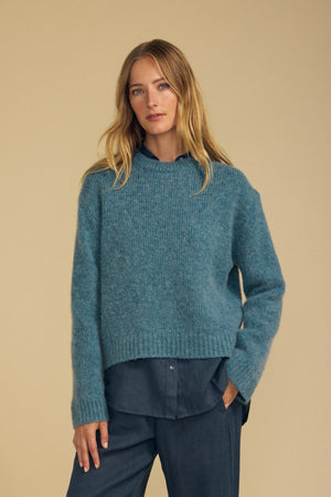 A person with long blonde hair wears the JUNE SWEATER by Velvet by Graham & Spencer—a cozy wool-cashmere piece in teal—layered over a blue button-up shirt and dark trousers, standing against a beige background.