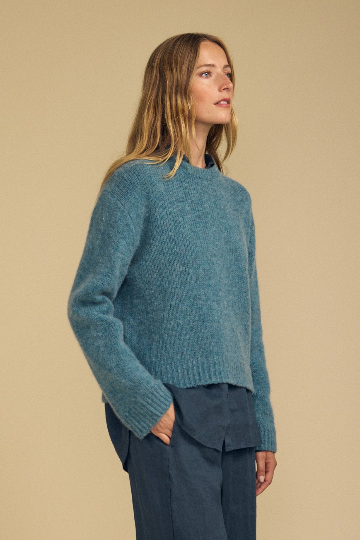 A person with long, light brown hair wears the JUNE SWEATER by Velvet by Graham & Spencer—a teal wool-cashmere knit with a modern silhouette—layered over a blue shirt and blue pants, standing against a plain beige background.