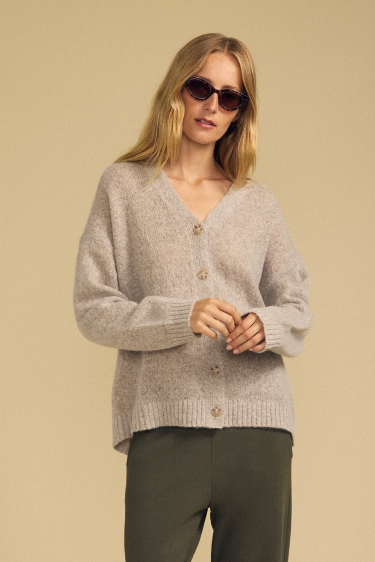 Wearing the LEA CARDIGAN by Velvet by Graham & Spencer in light gray wool-cashmere with olive green pants and sunglasses, this person stands against a plain beige background—an effortlessly stylish look perfect for everyday layering.-41964584796353
