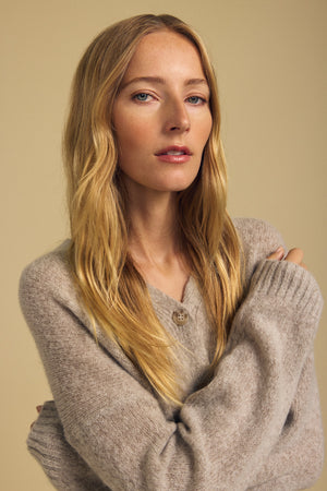 A person with long blond hair wearing the LEA CARDIGAN by Velvet by Graham & Spencer stands against a plain beige background, looking at the camera with a neutral expression—an effortless take on everyday layering.