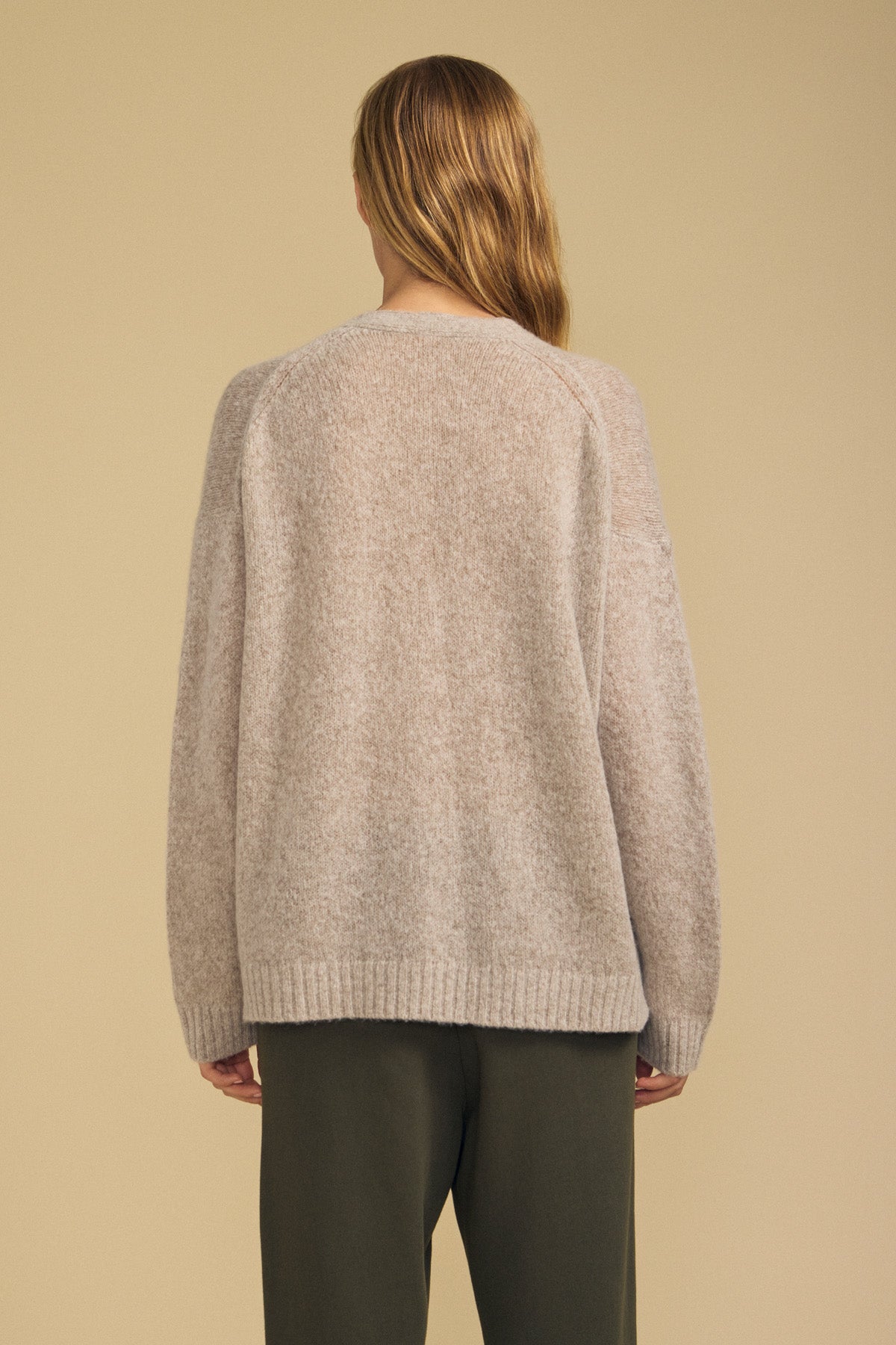 A person with long hair is seen from behind against a plain beige background, wearing the LEA CARDIGAN by Velvet by Graham & Spencer—a loose, beige knit sweater ideal for everyday layering—paired with dark pants.