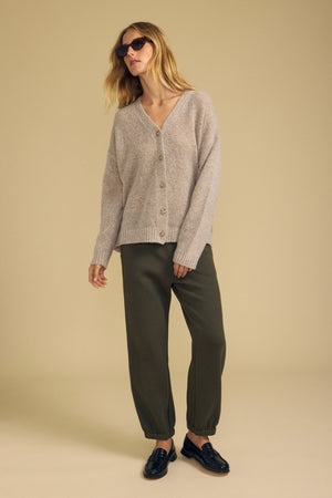 A woman wearing sunglasses, the LEA CARDIGAN by Velvet by Graham & Spencer in a cozy wool-cashmere blend in beige, paired with olive green jogger pants and black loafers, stands against a plain beige background—perfect for everyday layering.