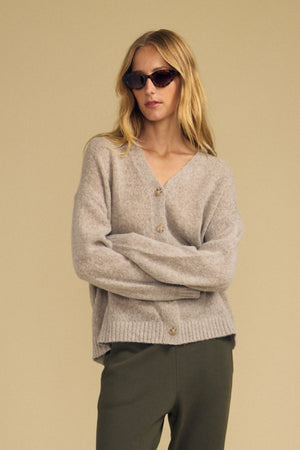 A woman wearing dark sunglasses, the LEA CARDIGAN by Velvet by Graham & Spencer—a beige wool-cashmere piece perfect for everyday layering—and olive green pants stands with her arms crossed against a plain beige background.