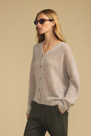 A person wearing sunglasses, the LEA CARDIGAN by Velvet by Graham & Spencer in light grey, and dark green pants—perfect for everyday layering—stands against a plain beige background.