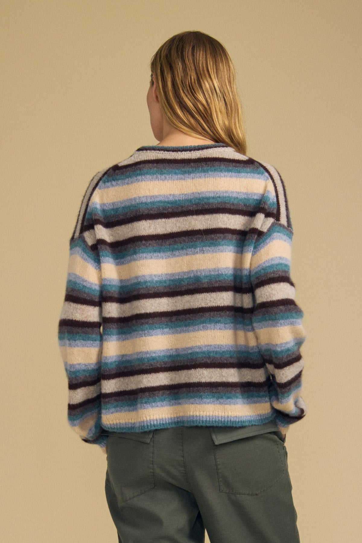 A person stands with their back to the camera, wearing the MAE SWEATER by Velvet by Graham & Spencer—a cozy wool-cashmere piece featuring blue, brown, and white stripes—paired with green pants against a plain beige background.