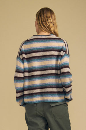 A person stands with their back to the camera, wearing the MAE SWEATER by Velvet by Graham & Spencer—a cozy wool-cashmere piece featuring blue, brown, and white stripes—paired with green pants against a plain beige background.