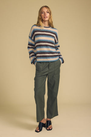 The woman wears the MAE SWEATER by Velvet by Graham & Spencer, paired with olive green pants and black slide sandals, standing with her hands in her pockets against a beige background.