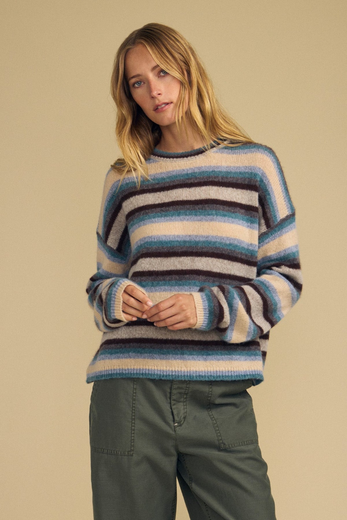 A woman stands against a beige background, wearing the MAE SWEATER by Velvet by Graham & Spencer—a cozy wool-cashmere striped sweater in shades of blue, brown, and beige—paired with green wide-leg pants.