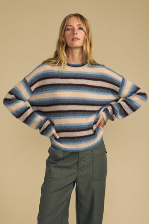 A woman stands against a beige background wearing the MAE SWEATER by Velvet by Graham & Spencer—a cozy wool-cashmere striped sweater in blue, cream, and brown tones—paired with olive green pants. Her hands rest on her hips as she looks directly at the camera.
