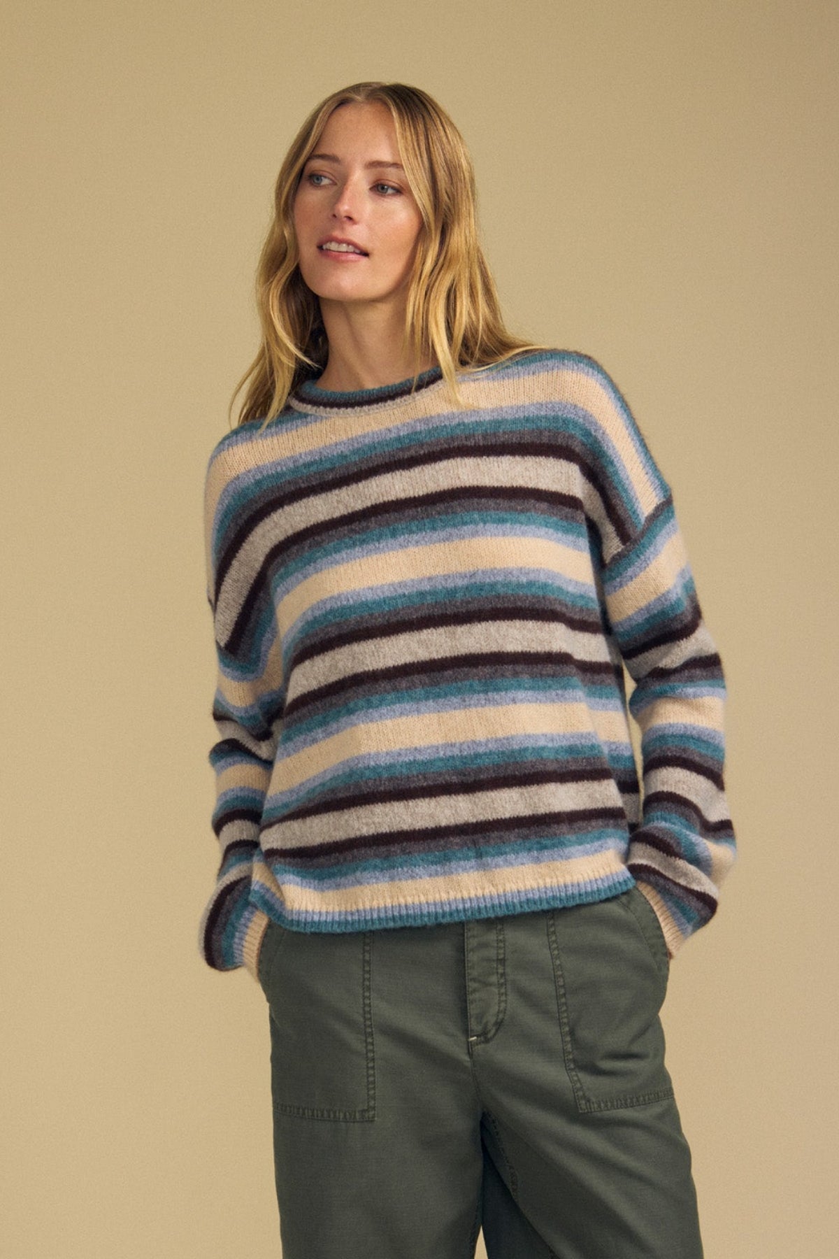 A person wearing the MAE SWEATER by Velvet by Graham & Spencer, featuring cozy blue, beige, and brown stripes, paired with green cargo pants and standing against a plain beige background.-41964639027393