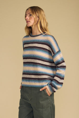 A person wearing the MAE SWEATER by Velvet by Graham & Spencer—a cozy wool-cashmere striped sweater in shades of blue, brown, and cream—paired with olive green pants stands against a beige background.