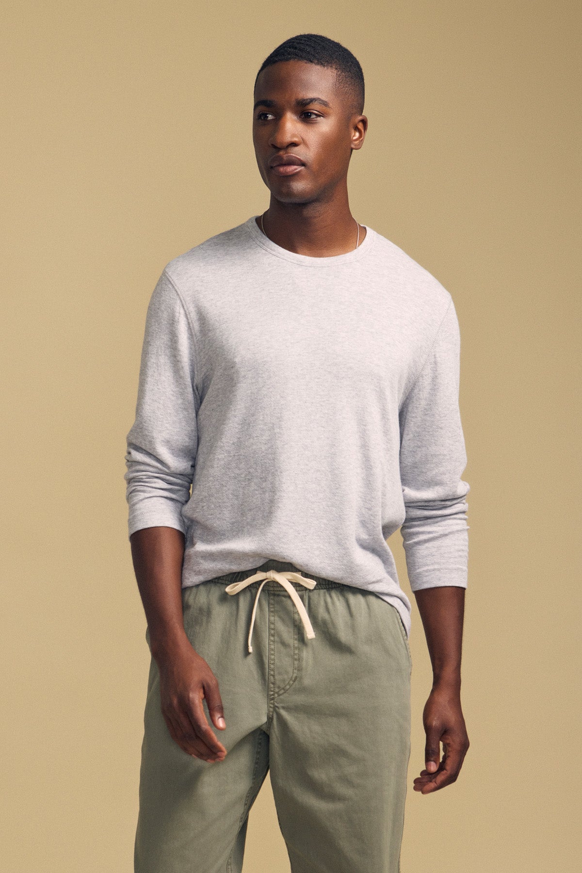 A man wearing the COLTON CREW by Velvet by Graham & Spencer—a light gray double-face jersey long-sleeve with a classic crew neckline—paired with olive green drawstring pants, stands against a plain beige background.-41859880452289