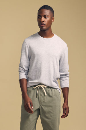 A man wearing the COLTON CREW by Velvet by Graham & Spencer—a light gray double-face jersey long-sleeve with a classic crew neckline—paired with olive green drawstring pants, stands against a plain beige background.