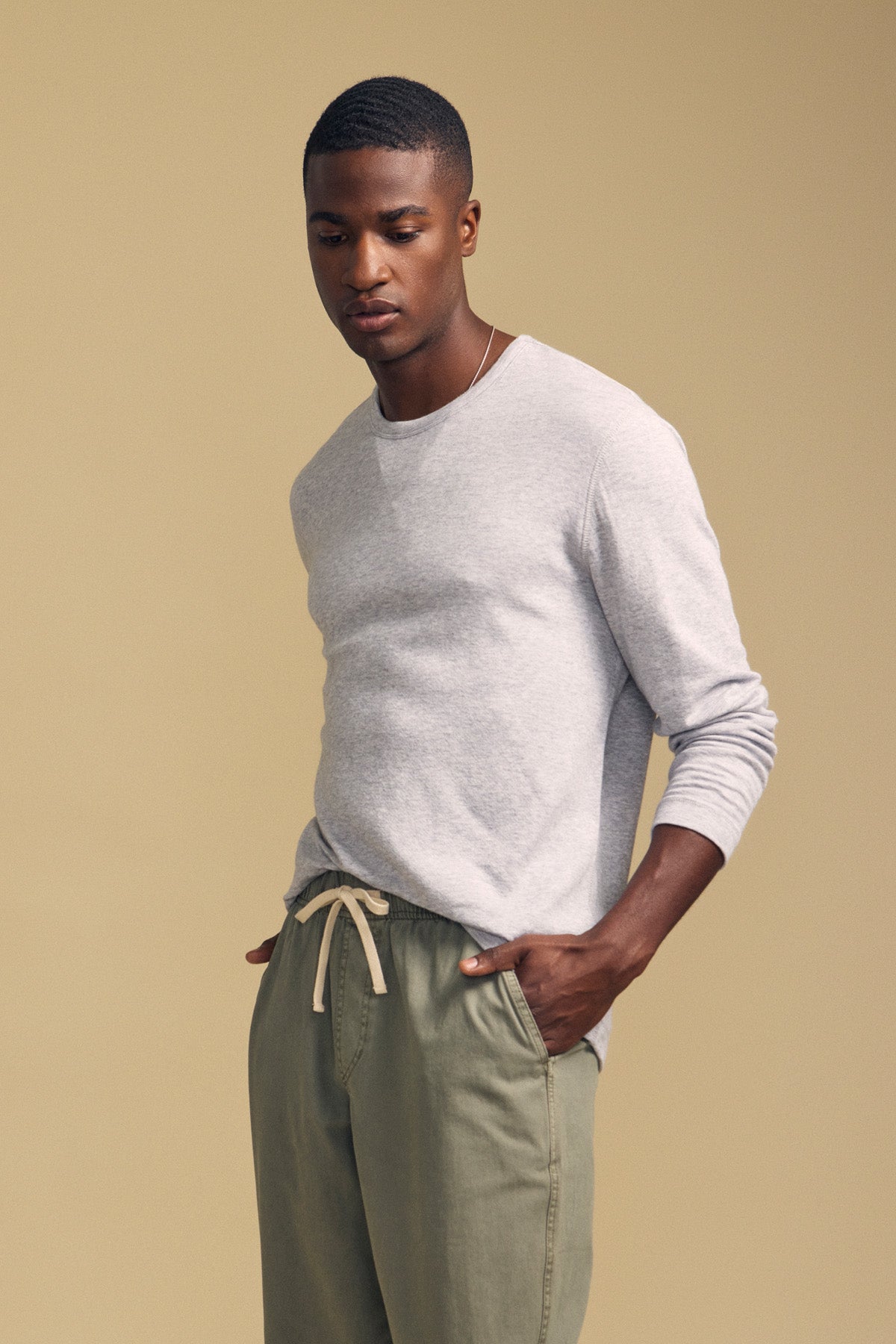 A man stands against a beige background, wearing the Velvet by Graham & Spencer COLTON CREW—a light gray long-sleeve top with a classic crew neckline—paired with olive green drawstring pants. His hands are casually in his pockets as he looks down.