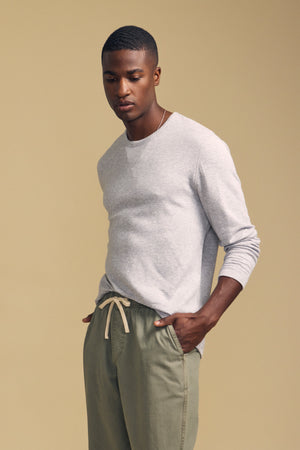 A man stands against a beige background, wearing the Velvet by Graham & Spencer COLTON CREW—a light gray long-sleeve top with a classic crew neckline—paired with olive green drawstring pants. His hands are casually in his pockets as he looks down.