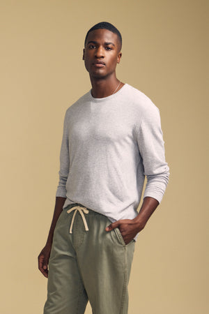 A person stands against a beige background, wearing the Velvet by Graham & Spencer COLTON CREW—a light gray long-sleeve crew shirt with a classic crew neckline—paired with olive green drawstring pants and one hand in their pocket.