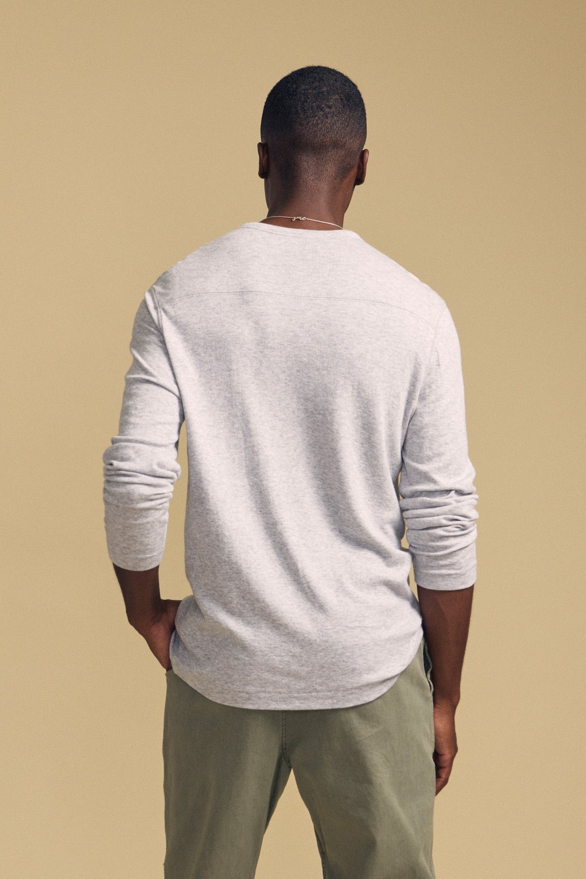 A person stands facing away, wearing the Velvet by Graham & Spencer COLTON CREW in light gray—a classic long-sleeve crewneck shirt—paired with olive green pants against a beige background.