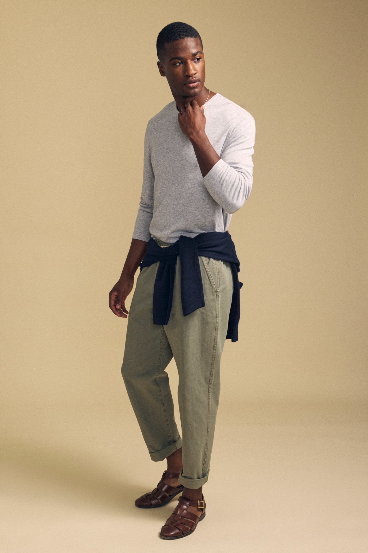 A man stands against a beige background wearing the Velvet by Graham & Spencer COLTON CREW, a light gray long-sleeve crew shirt with a classic neckline, paired with olive green pants, brown sandals, and a dark sweater tied around his waist.
