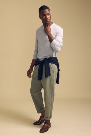A man stands against a beige background wearing the Velvet by Graham & Spencer COLTON CREW, a light gray long-sleeve crew shirt with a classic neckline, paired with olive green pants, brown sandals, and a dark sweater tied around his waist.