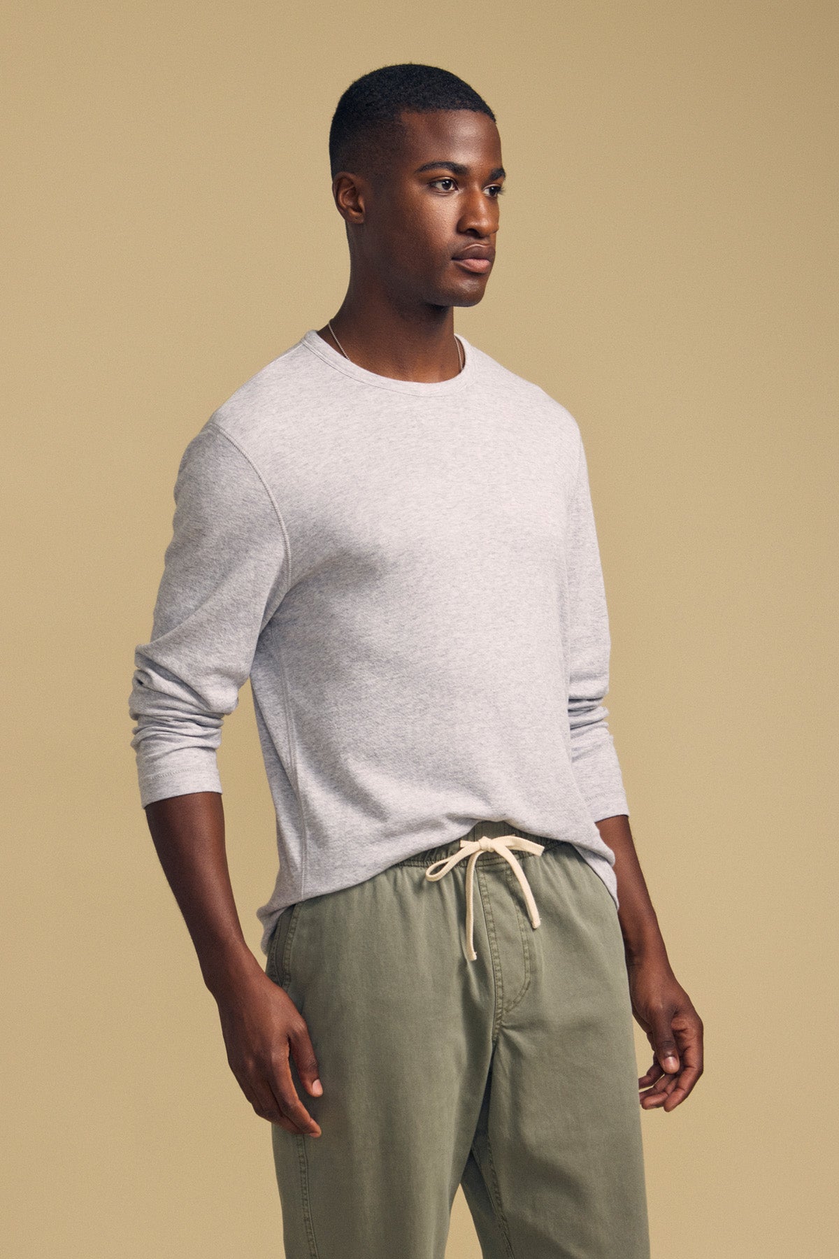 A man stands against a beige background, wearing the Velvet by Graham & Spencer COLTON CREW, a light gray long-sleeve top crafted from soft double-face jersey, paired with olive green drawstring pants.